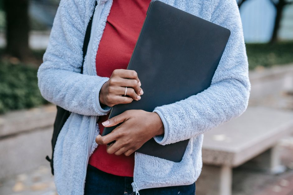 Crop anonymous young African American female student in warm casual clothes and with backpack standing in campus park with laptop in hands
